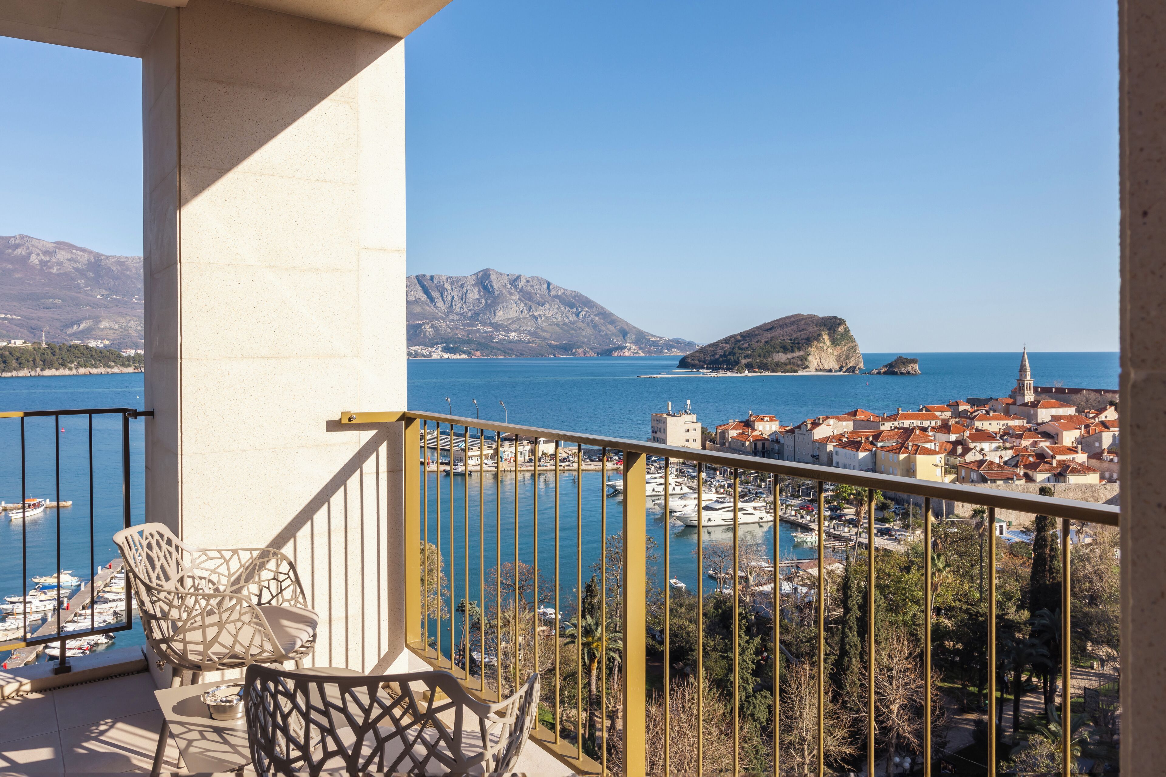 A balcony with two chairs and a table, overlooking a coastal town with boats and a mountain in the background.