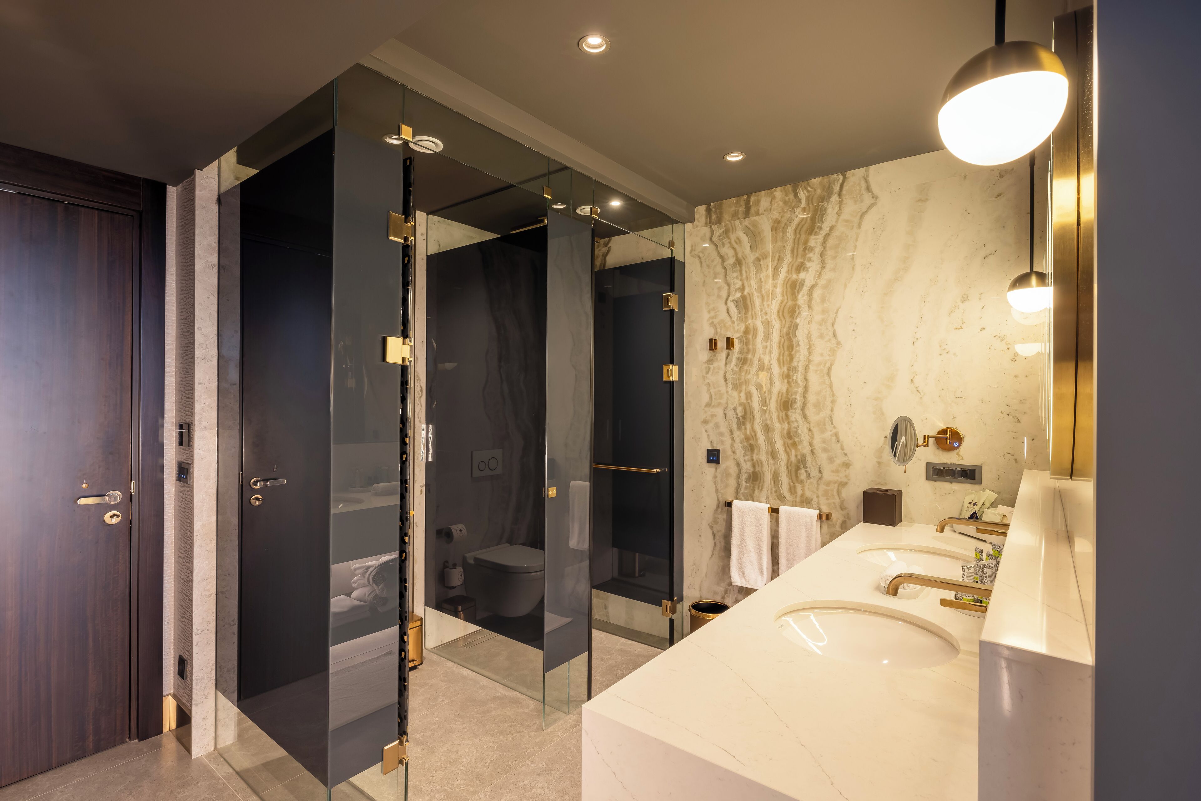 Un baño moderno con ducha de vidrio, pared de mármol y tocador con doble lavamanos.