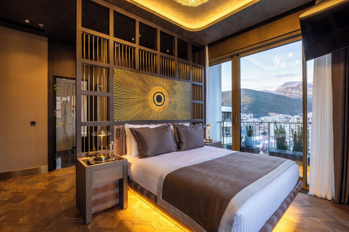 Une chambre moderne avec un grand lit, une table de chevet, un panneau décoratif au mur et une vue sur la ville grâce à de grandes fenêtres.