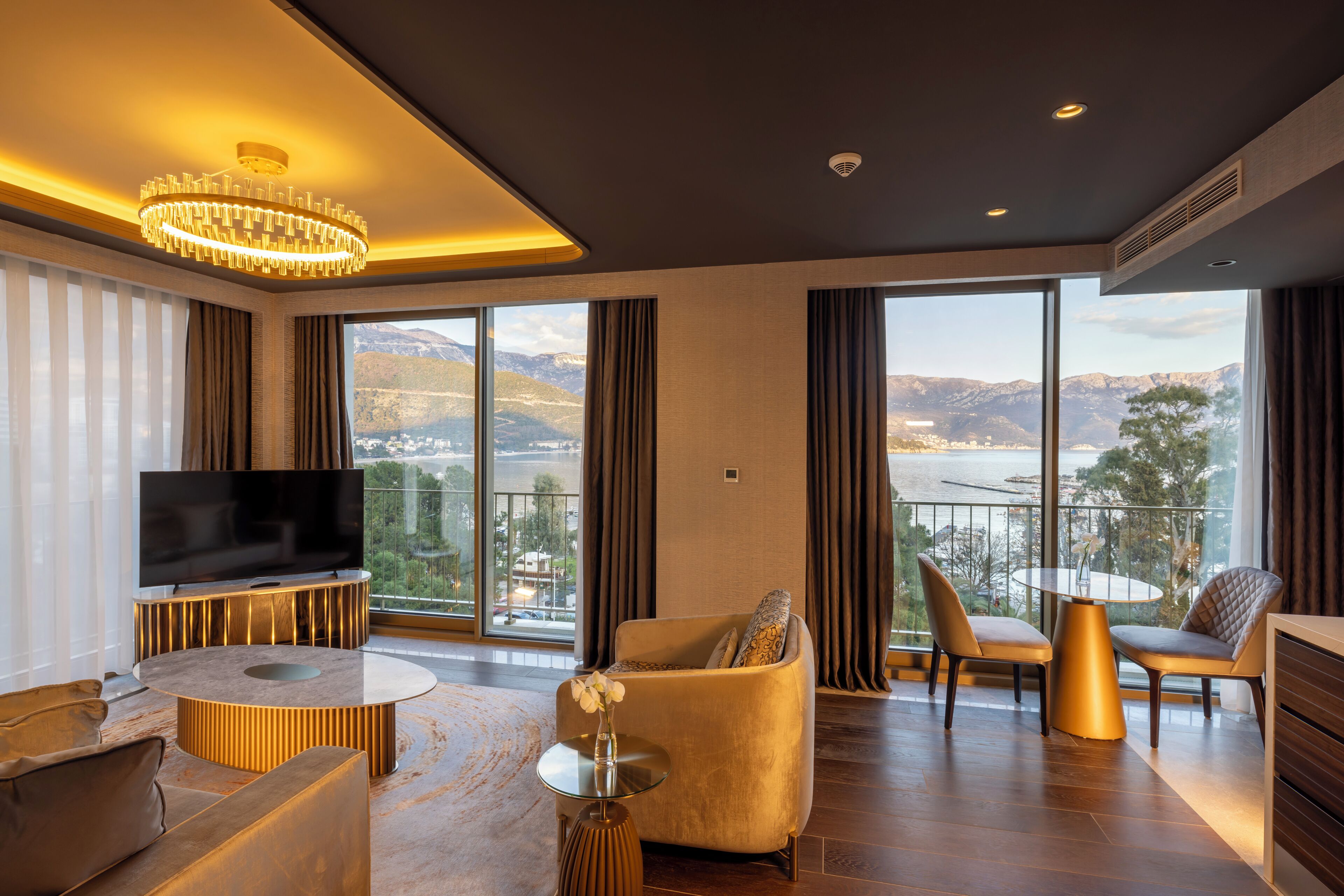 A modern living room with a large TV, comfortable seating, a round coffee table, and a balcony with a scenic view.