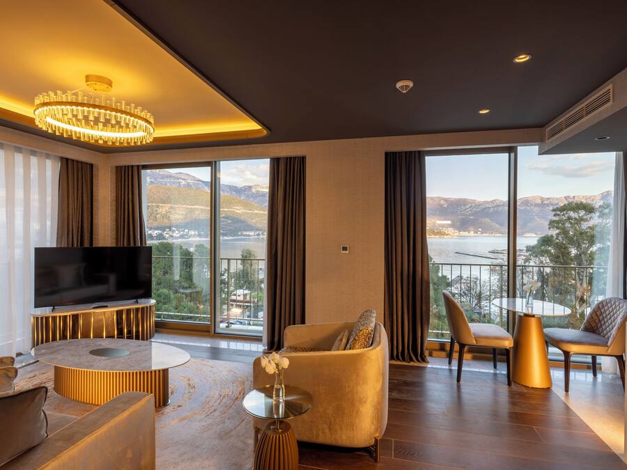 A modern living room with a large TV, comfortable seating, a round coffee table, and a balcony with a scenic view.