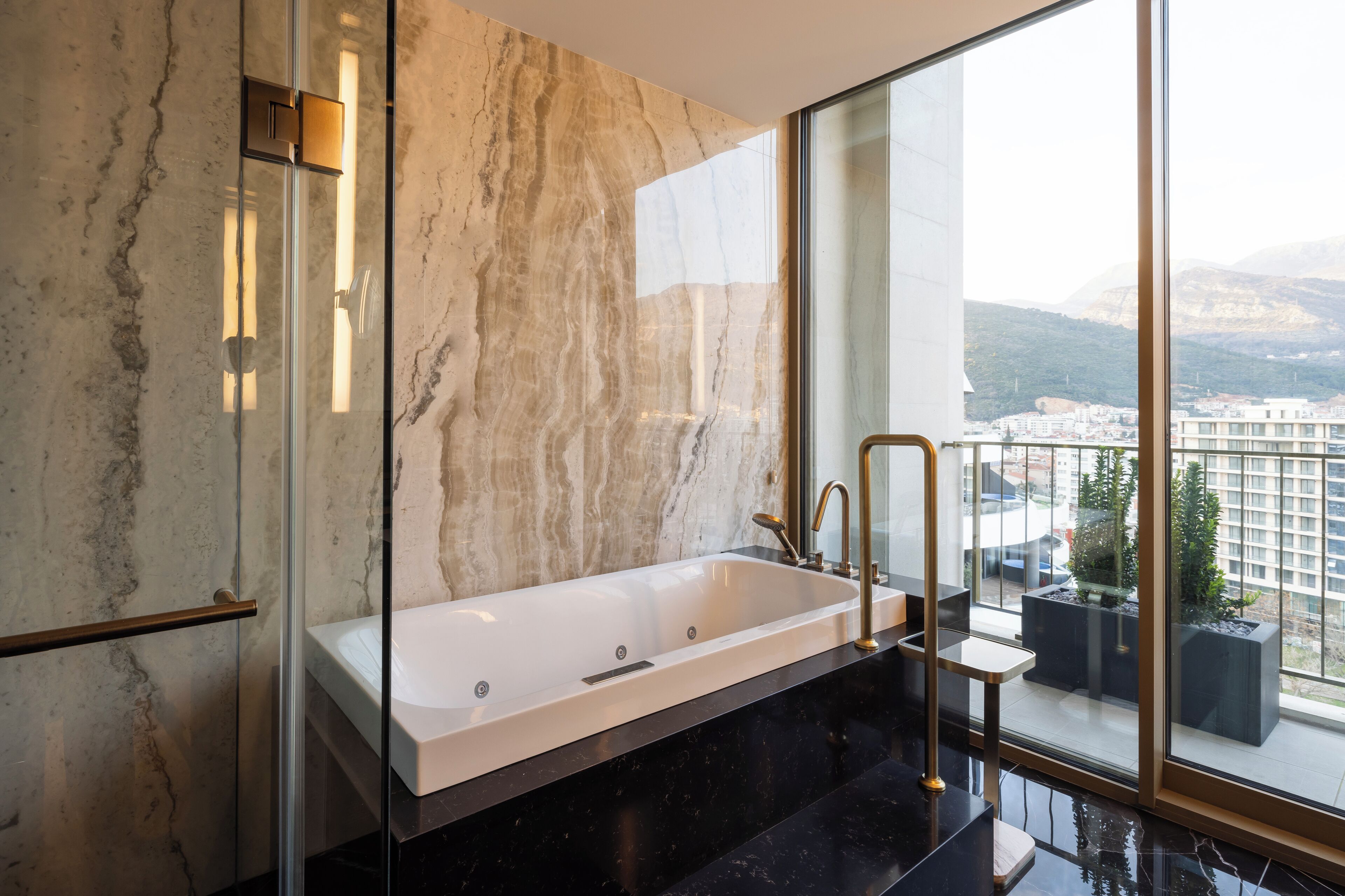 A modern bathroom with a large bathtub, a glass-enclosed shower, and a view of a cityscape through large windows.