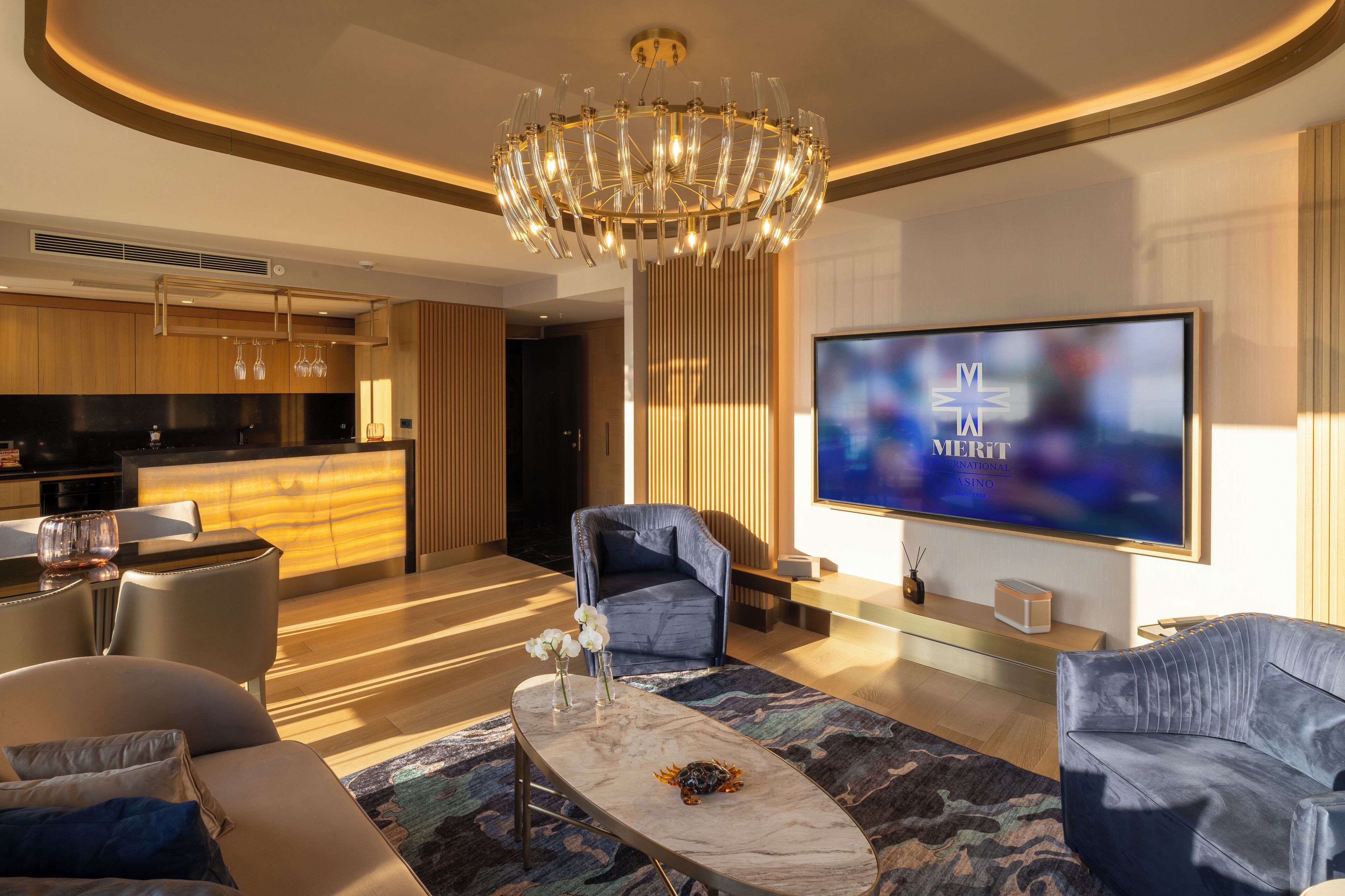 Una sala de estar moderna con un televisor de pantalla plana grande, una araña y una cocina al fondo.