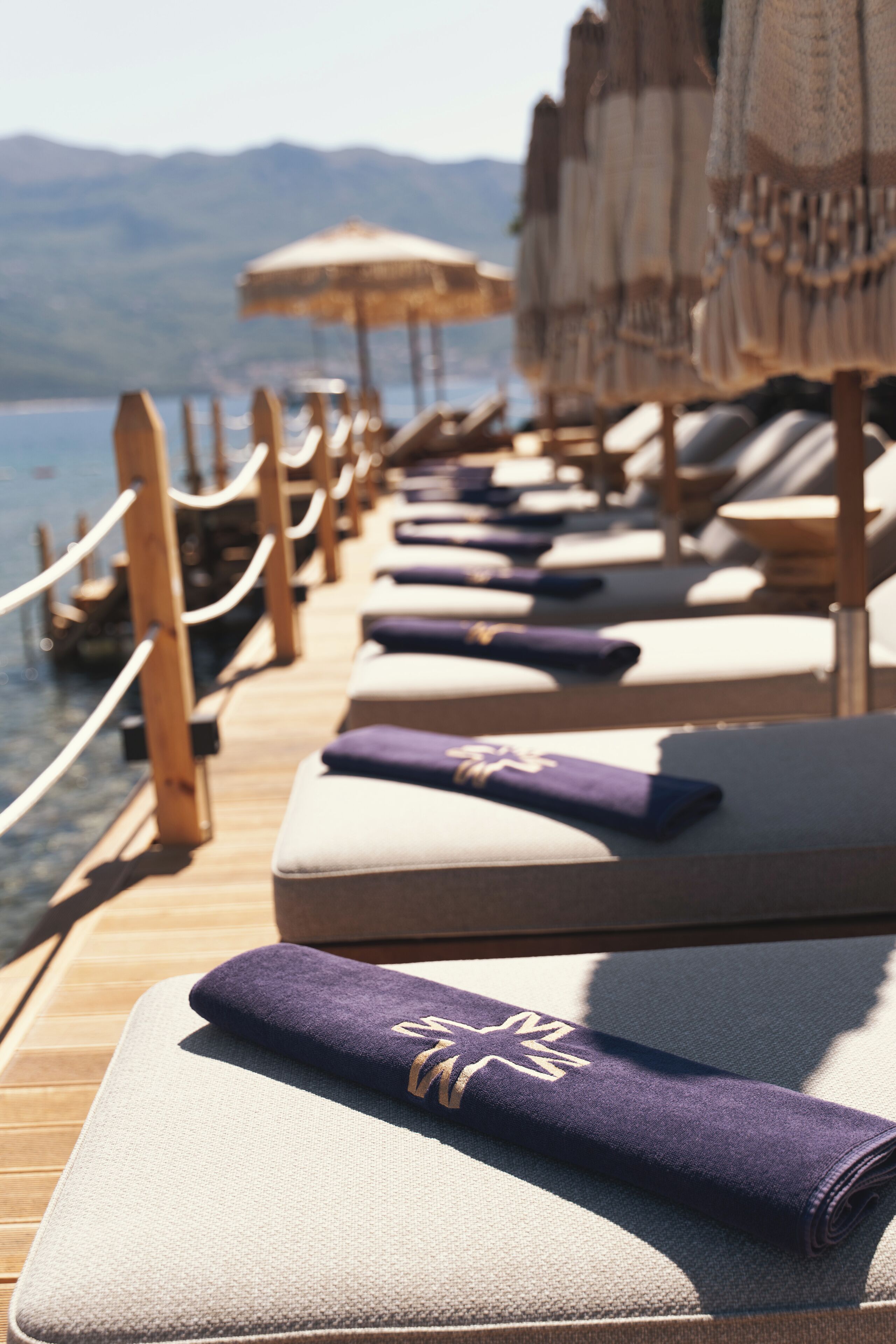 A row of sun loungers with towels, wooden deck, and umbrellas, overlooking a body of water with mountains in the background.