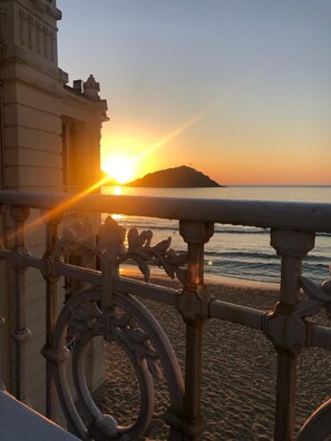 Une vue sur le coucher de soleil depuis un balcon orné de balustrades en métal ouvragé, donnant sur une plage et une île lointaine.