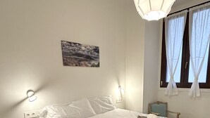 A neatly made bed with a white and brown blanket, a bedside table, a wall-mounted lamp, and a painting on the wall. - Corazón Donostiarra (Donostia)