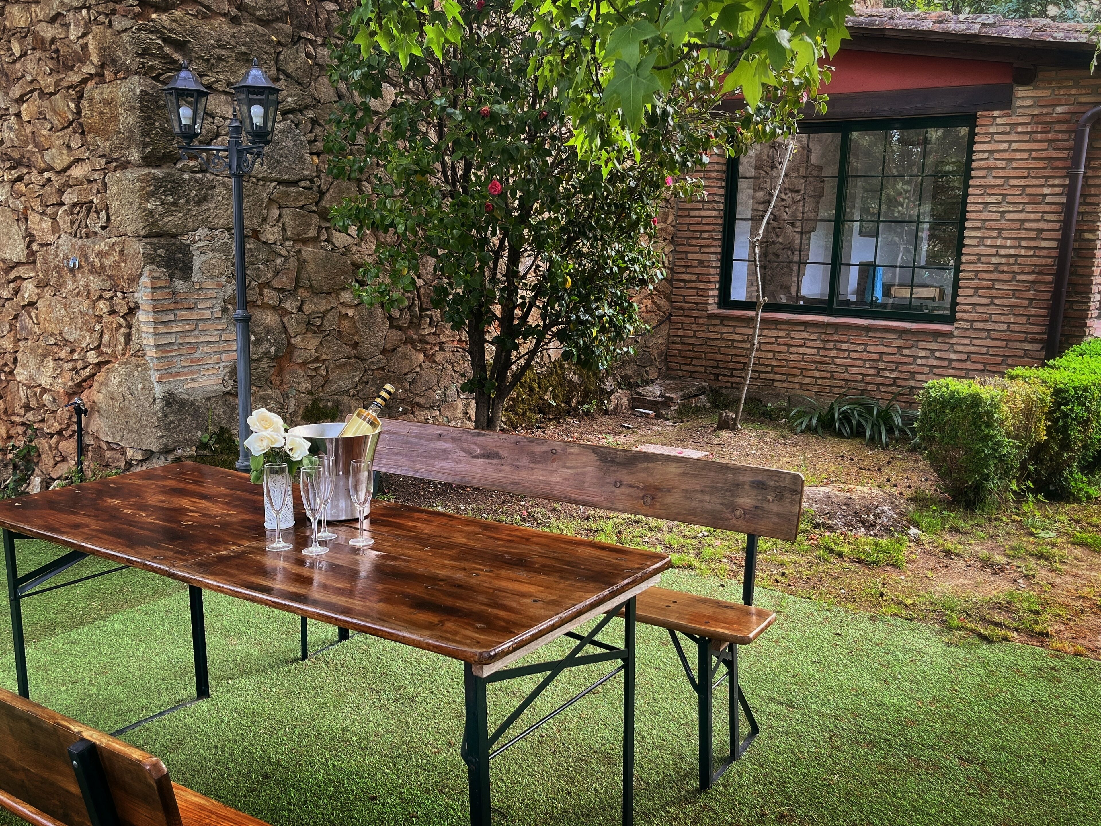 A wooden table with a bottle of champagne and glasses, set against a stone wall with a bench and a tree.