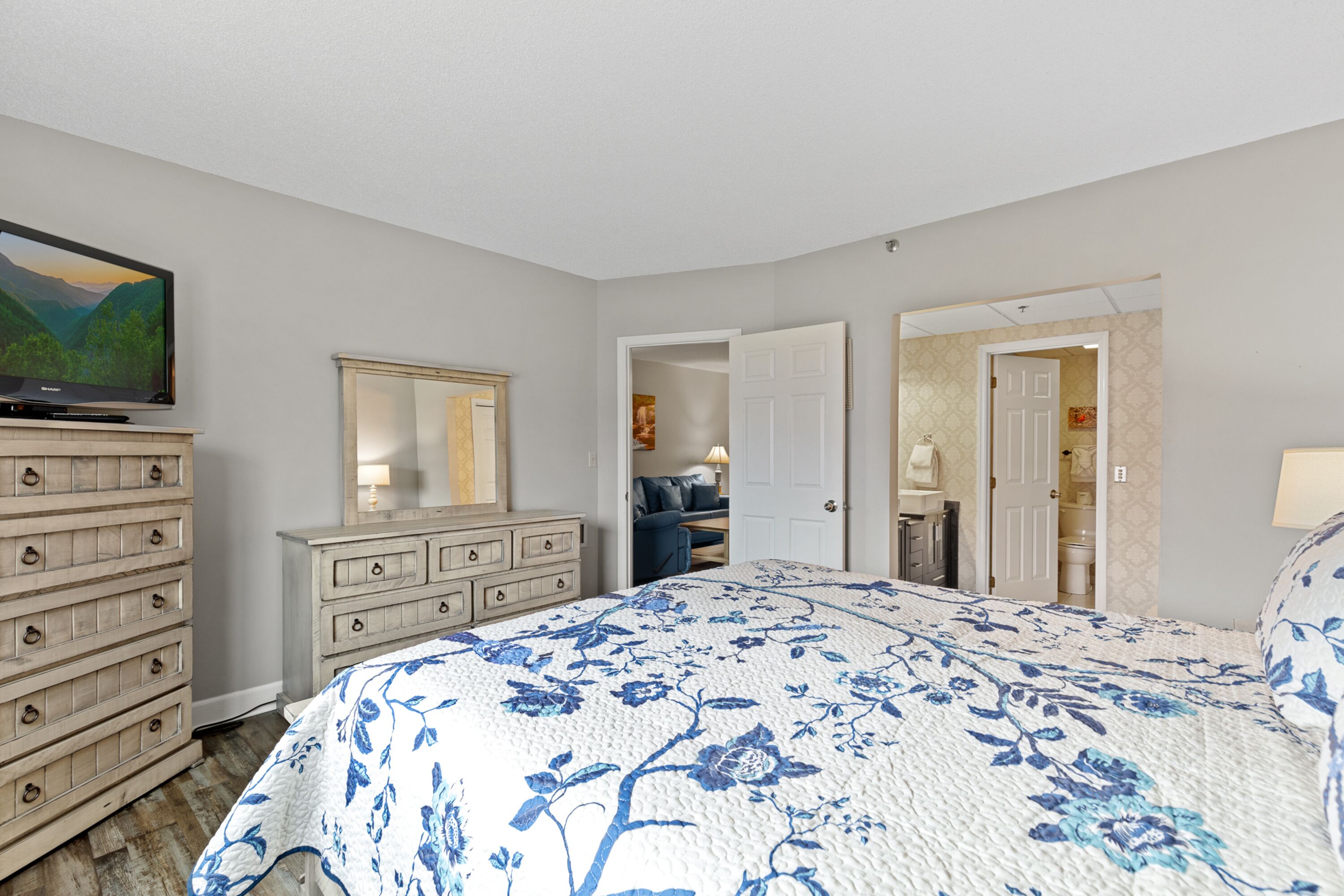 A bedroom with a large bed, a dresser, a television, and a door leading to another room.