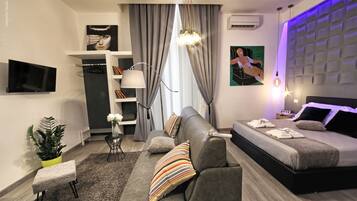 A modern bedroom with a sofa, a bed, a TV, a plant, and a window with curtains.