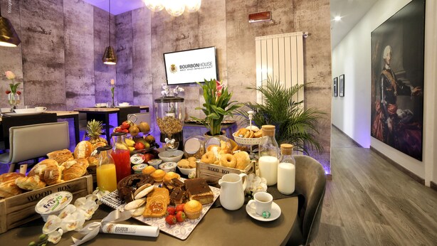 A breakfast buffet with various pastries, fruits, and beverages on a wooden table in a modern room with a chandelier and artwork on the walls. - Bourbon House (Naples)