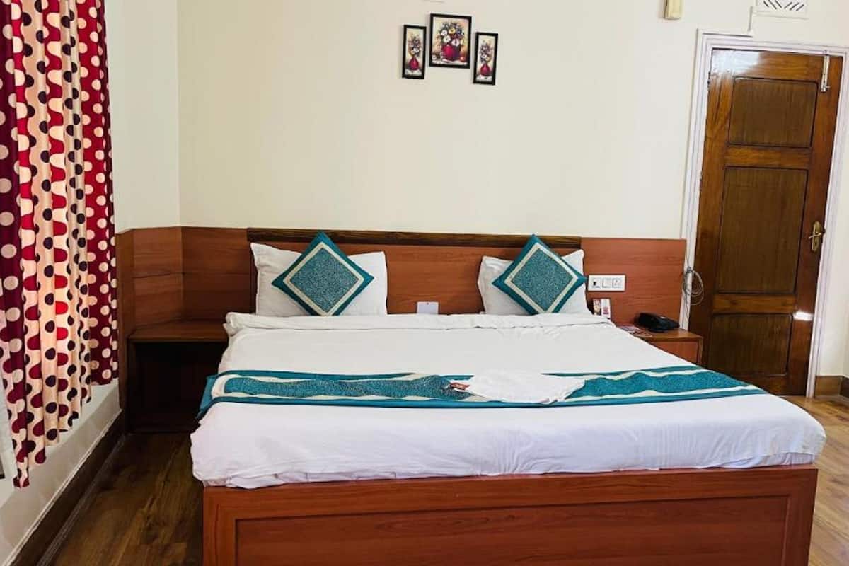 A neatly made bed with a white and teal bedspread, two matching pillows, and a wooden headboard. A wooden nightstand with a phone and a small lamp is beside the bed. A red and white polka dot curtain is on the left.