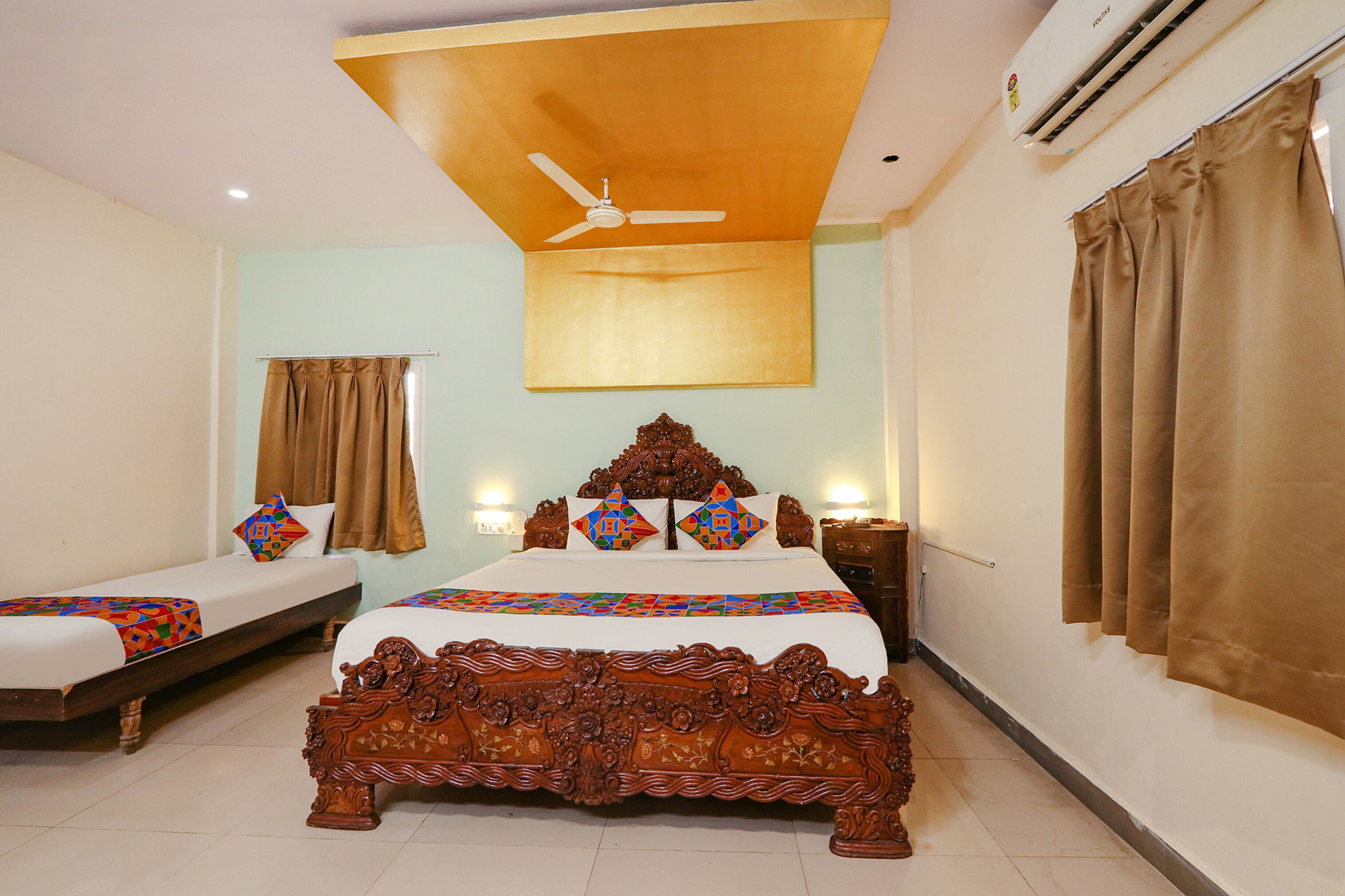 A hotel room with a large bed, a smaller bed, a ceiling fan, an air conditioner, and curtains.
