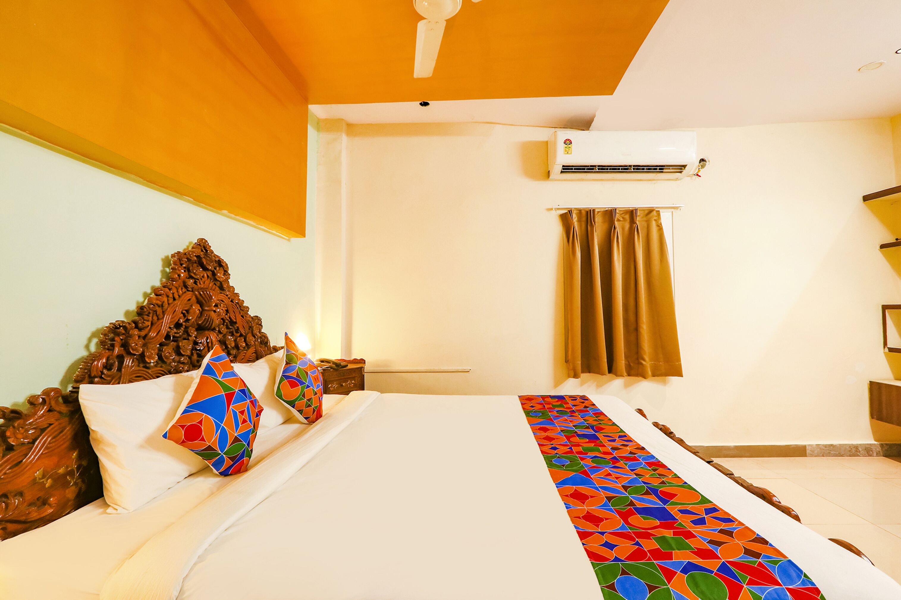 A hotel room with a bed, a ceiling fan, an air conditioning unit, a window with curtains, and a wooden headboard.