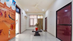 A hallway with a tiled floor, a ceiling fan, a wall-mounted air conditioner, a door with a red frame, and a decorative wall with a peacock and a figure. - Fabhotel Moon Light Palace (Jaipur)