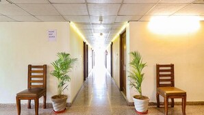 Um corredor com duas cadeiras de madeira e plantas em vasos, um aviso na parede e uma luz intensa no final.