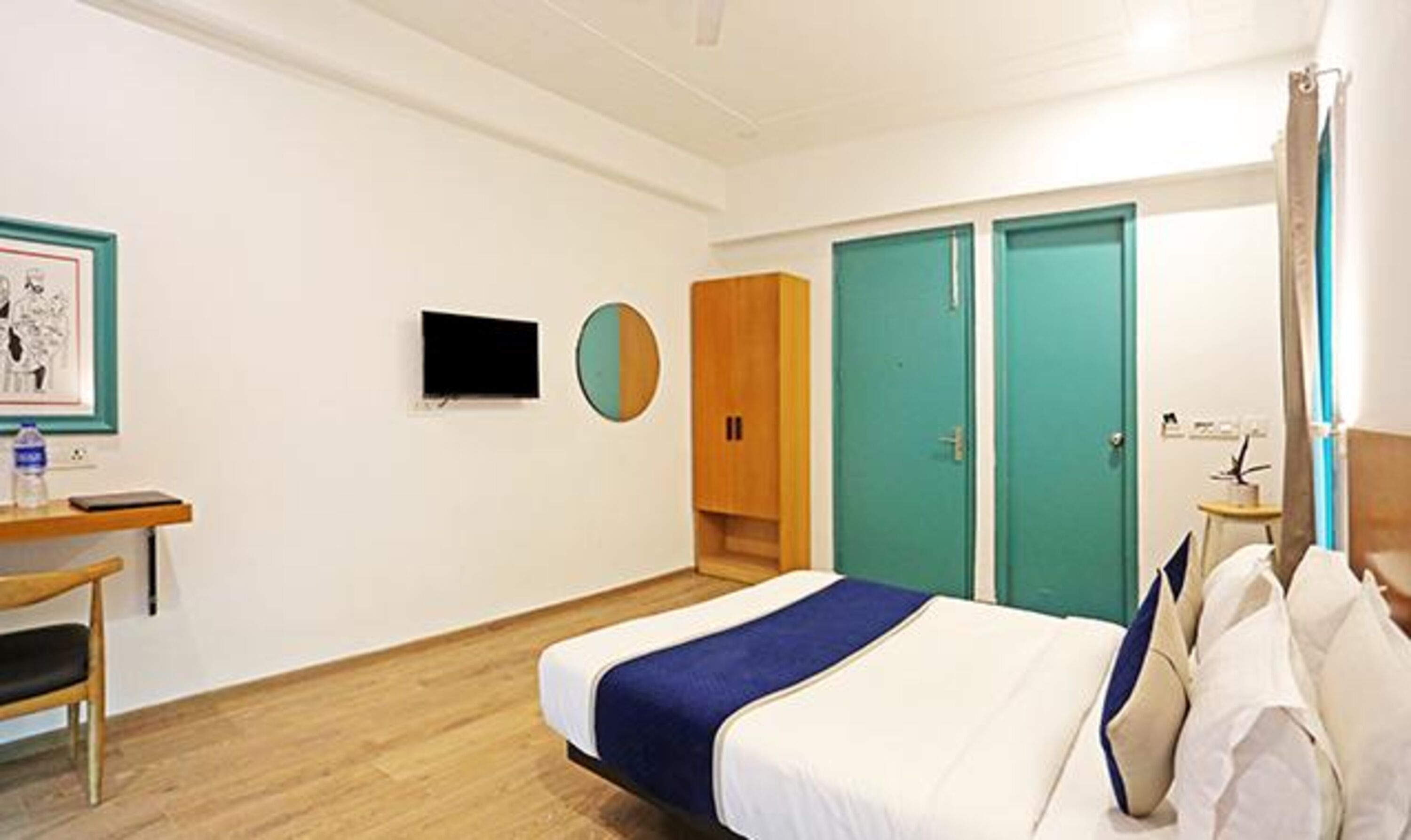 A hotel room with a bed, a desk with a chair, a TV mounted on the wall, and two doors.