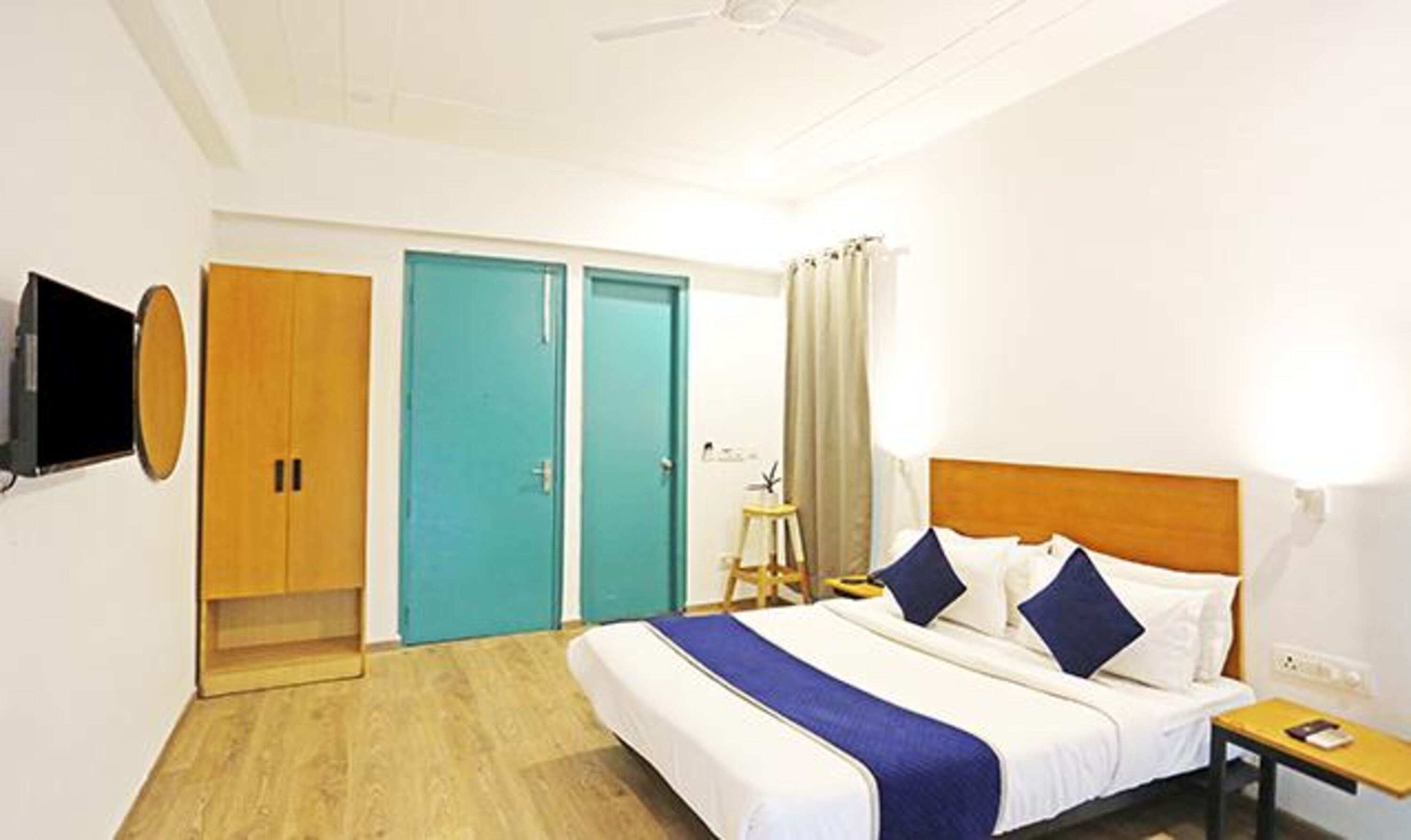 Ein Hotelzimmer mit Bett, Holzschrank, türkisgrüner Tür, kleinem Tisch und an der Wand montiertem Fernseher.