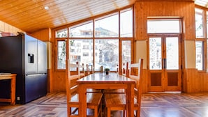 A wooden interior with a dining table and benches, a black refrigerator, and large windows with a view of buildings and trees. - Fabhotel Manali Heaven (Manali)