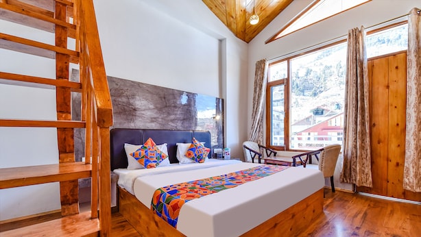 A wooden bed with a colorful blanket, a wooden chair, and a view of buildings through large windows. - Fabhotel Manali Heaven (Manali)