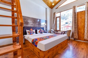 A wooden bed with a colorful blanket, a wooden chair, and a view of buildings through large windows. - Fabhotel Manali Heaven (Manali)