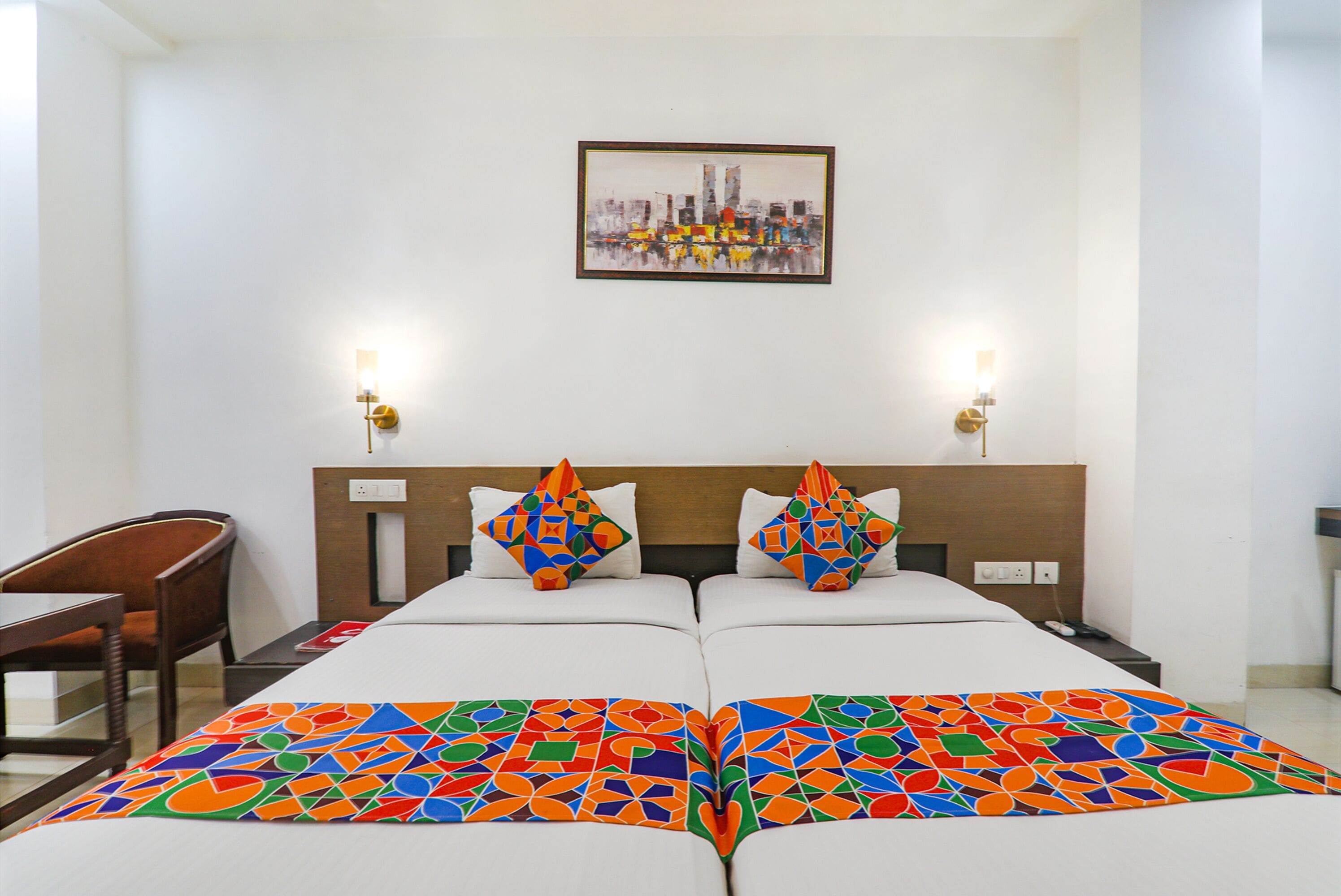 A hotel room with a bed, two bedside tables, a chair, a cityscape painting, and two wall-mounted lamps.