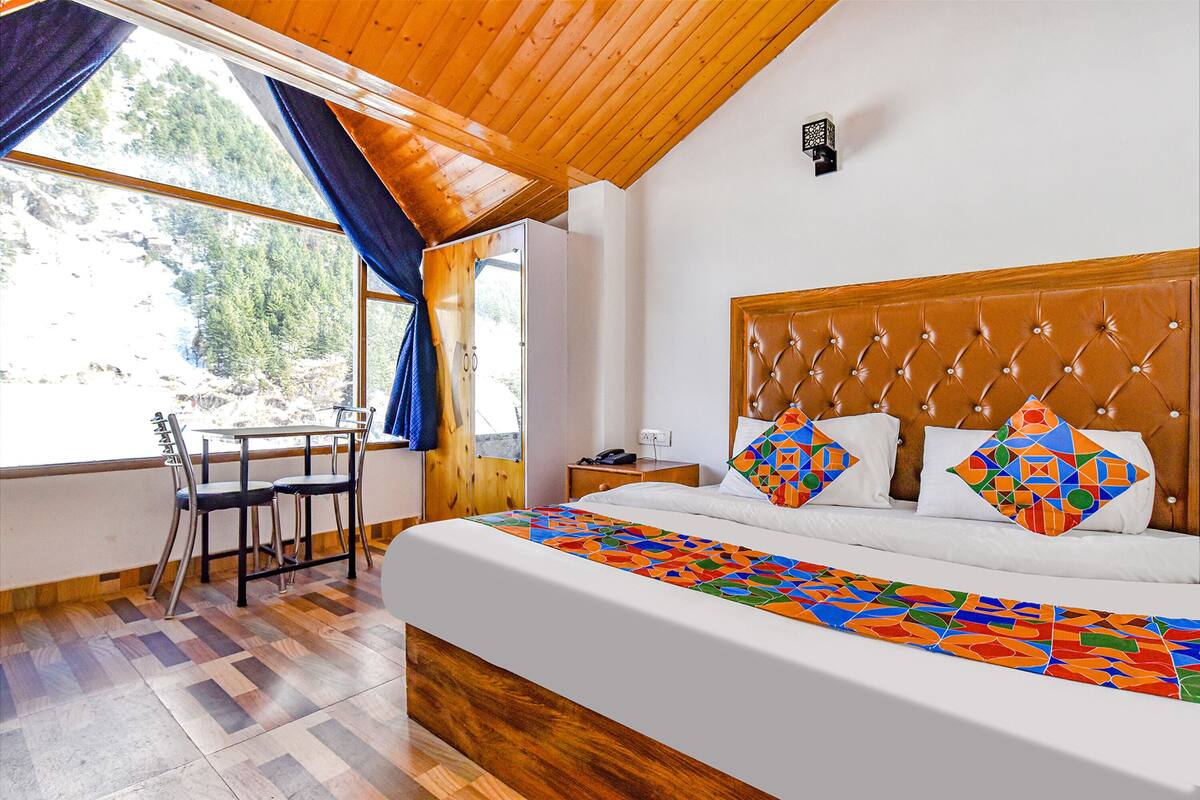 Quarto com uma cama grande, cabeceira de madeira, janela com cortinas, uma mesinha com cadeiras e um criado-mudo com telefone.