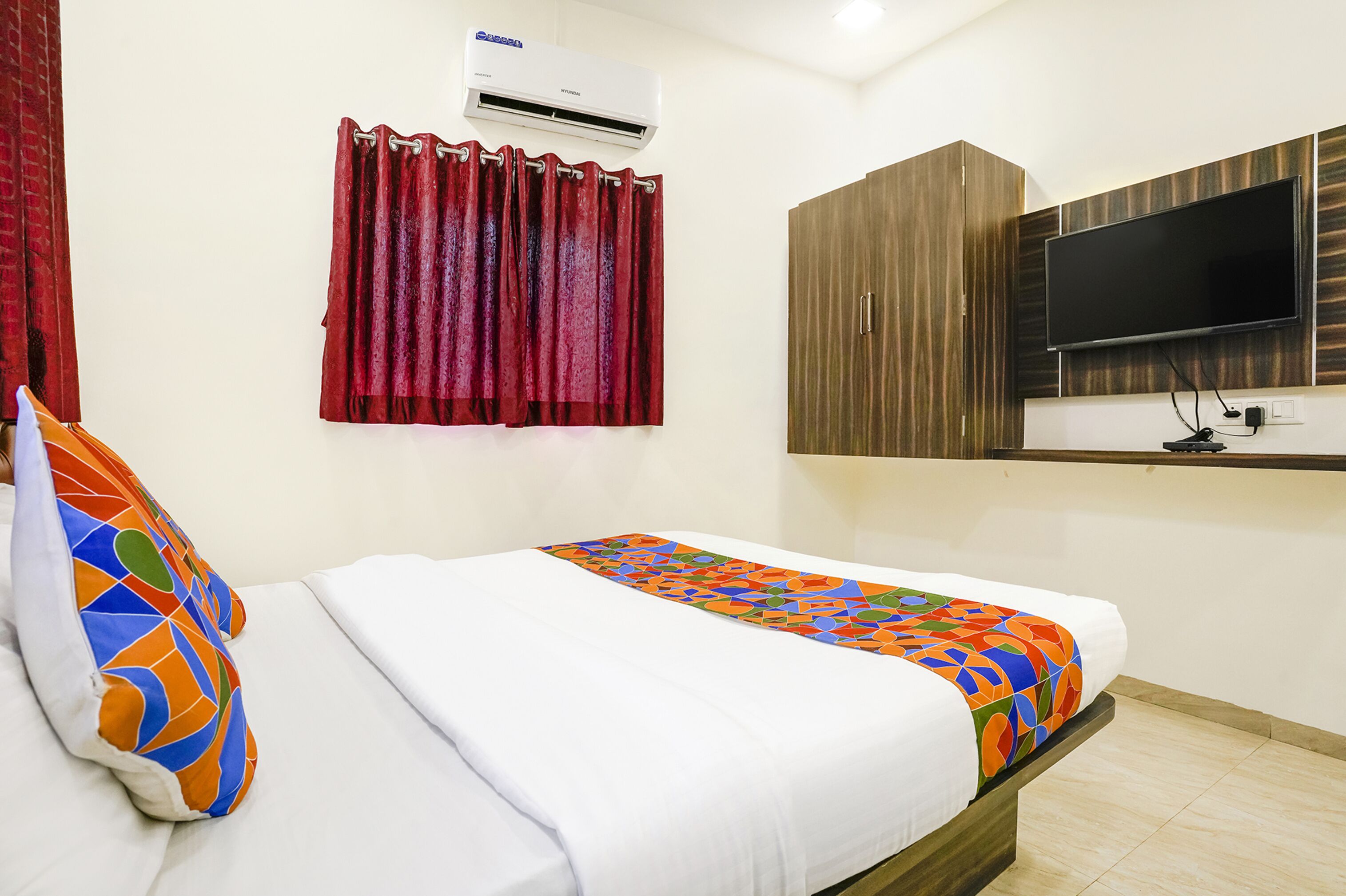 A neatly made bed with a colorful geometric-patterned blanket, a red curtain, a mounted TV, and a wooden wardrobe.