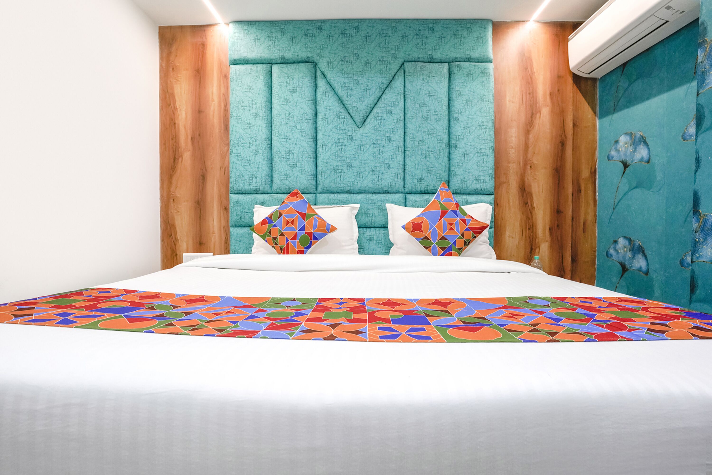 A modern hotel room with a teal headboard, a geometric-patterned bedspread, and a wooden accent wall.