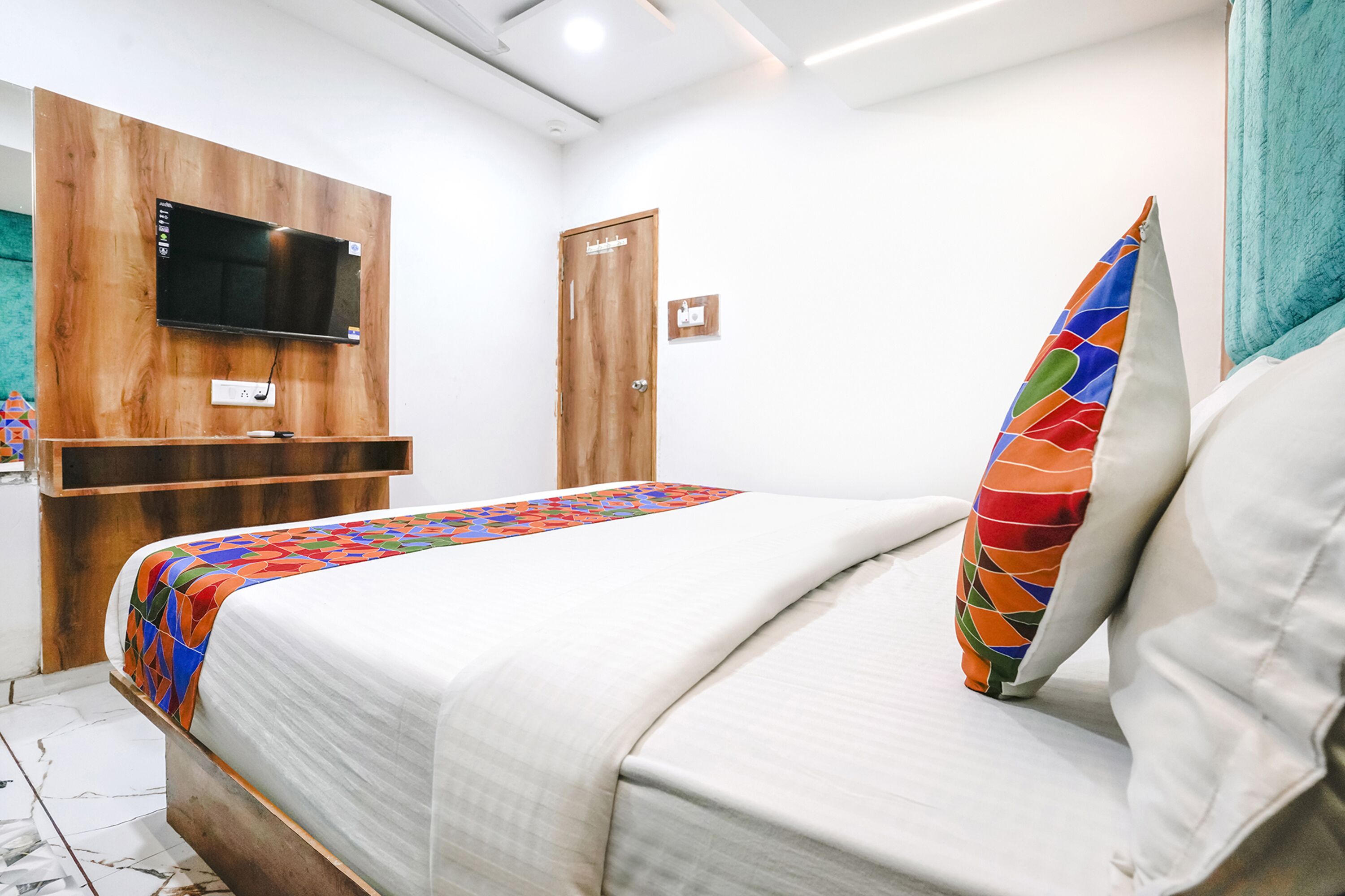 A bedroom with a bed, a TV mounted on a wooden panel, a door, and a colorful pillow.