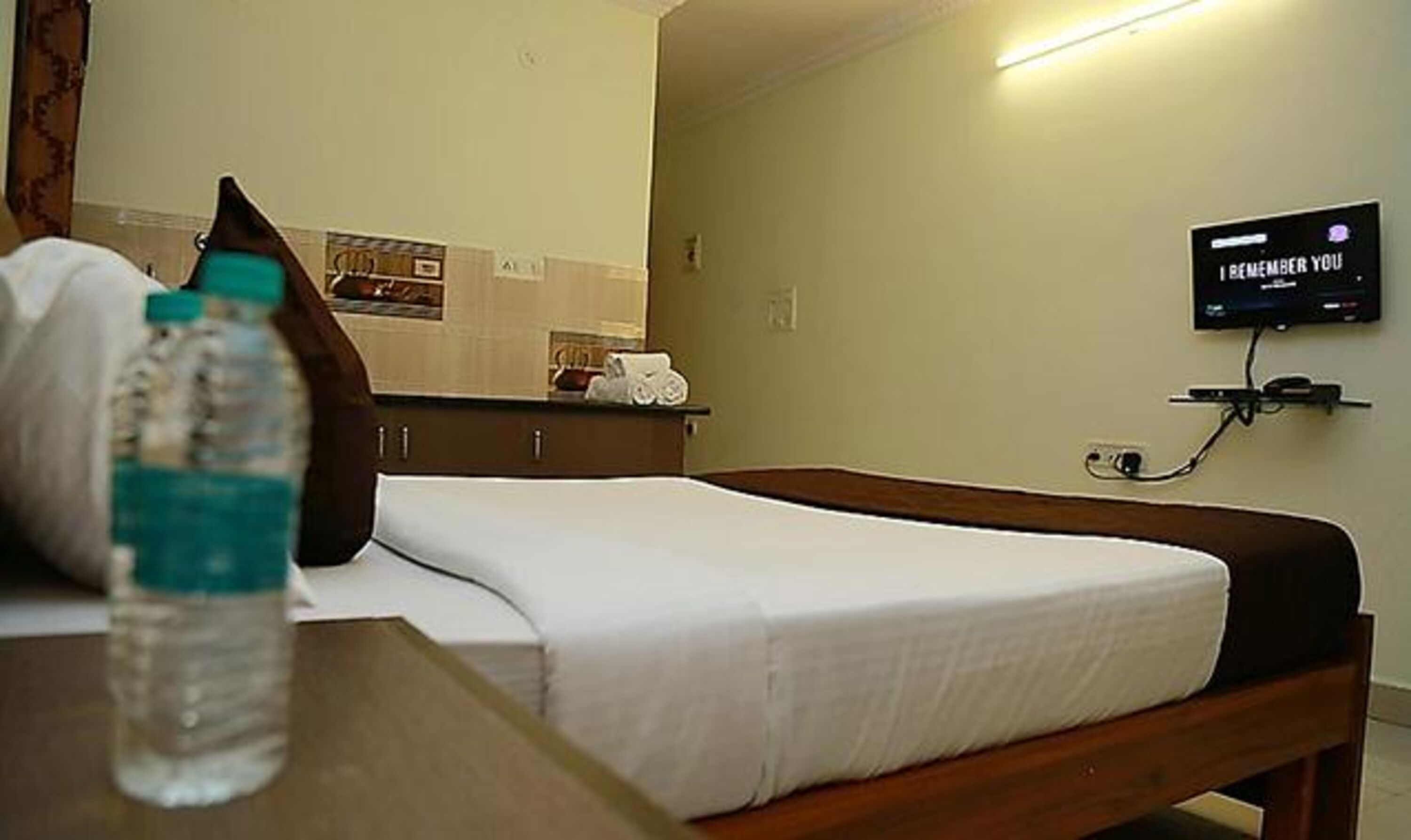 A hotel room with a bed, a bedside table with a water bottle, a TV mounted on the wall, and a mirror above the bed.