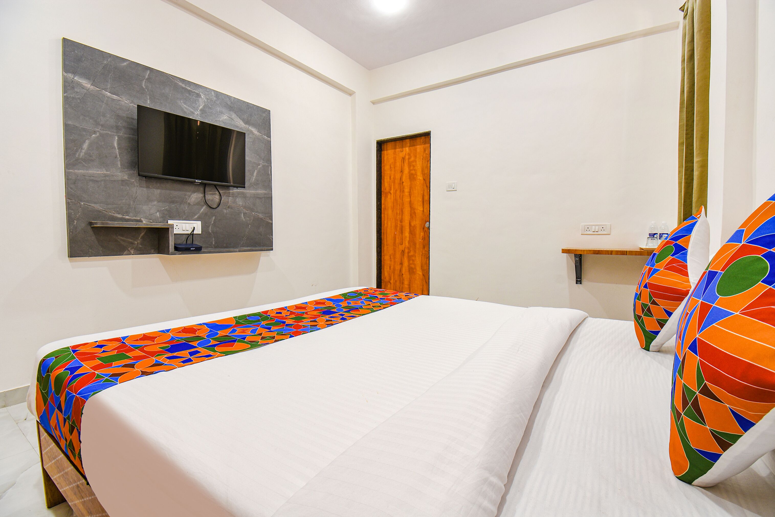 A hotel room with a bed, a TV mounted on the wall, a wooden door, and a small shelf with a bottle.