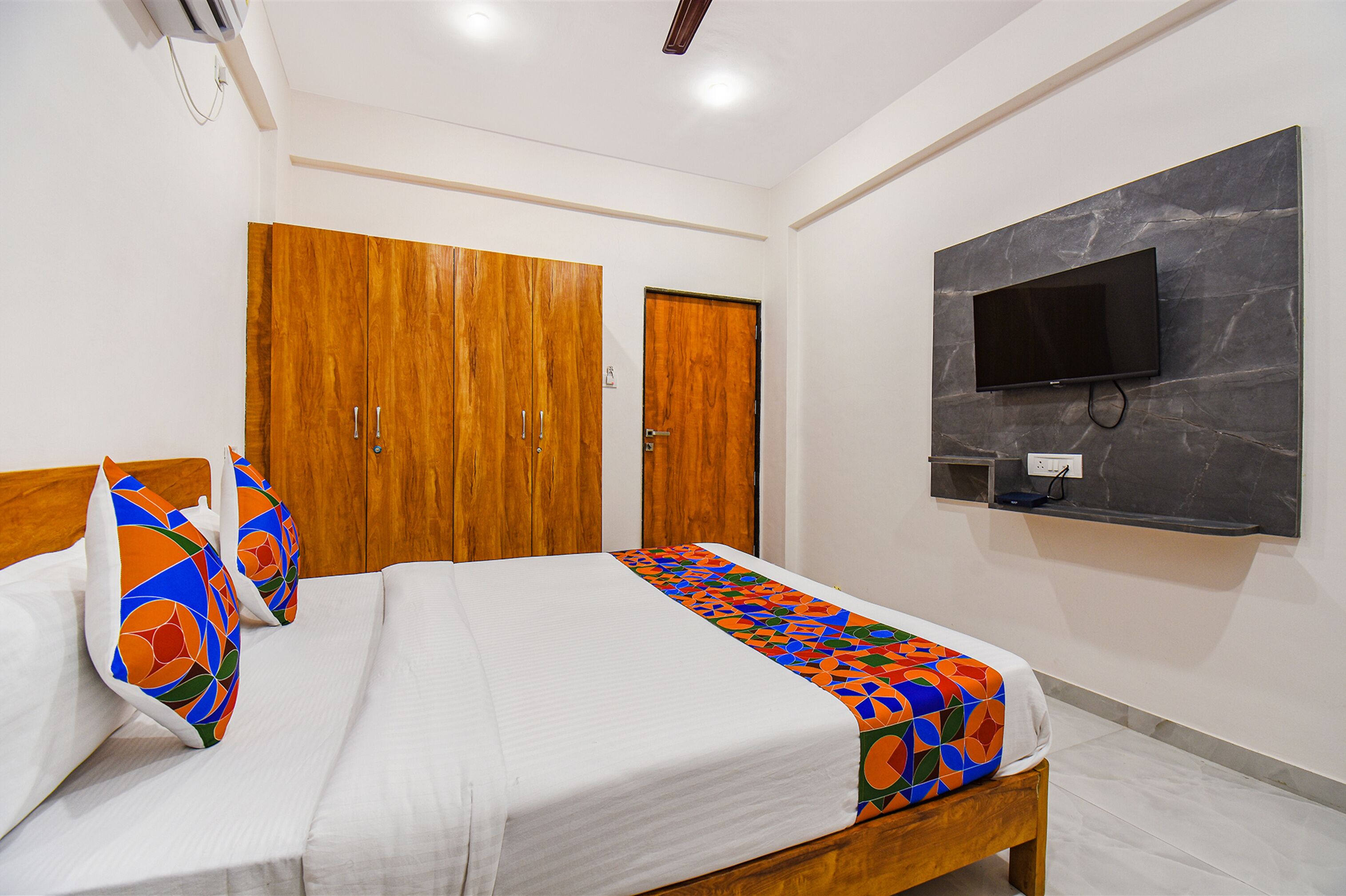 A hotel room with a bed, a TV mounted on the wall, wooden wardrobes, and a colorful bedspread.