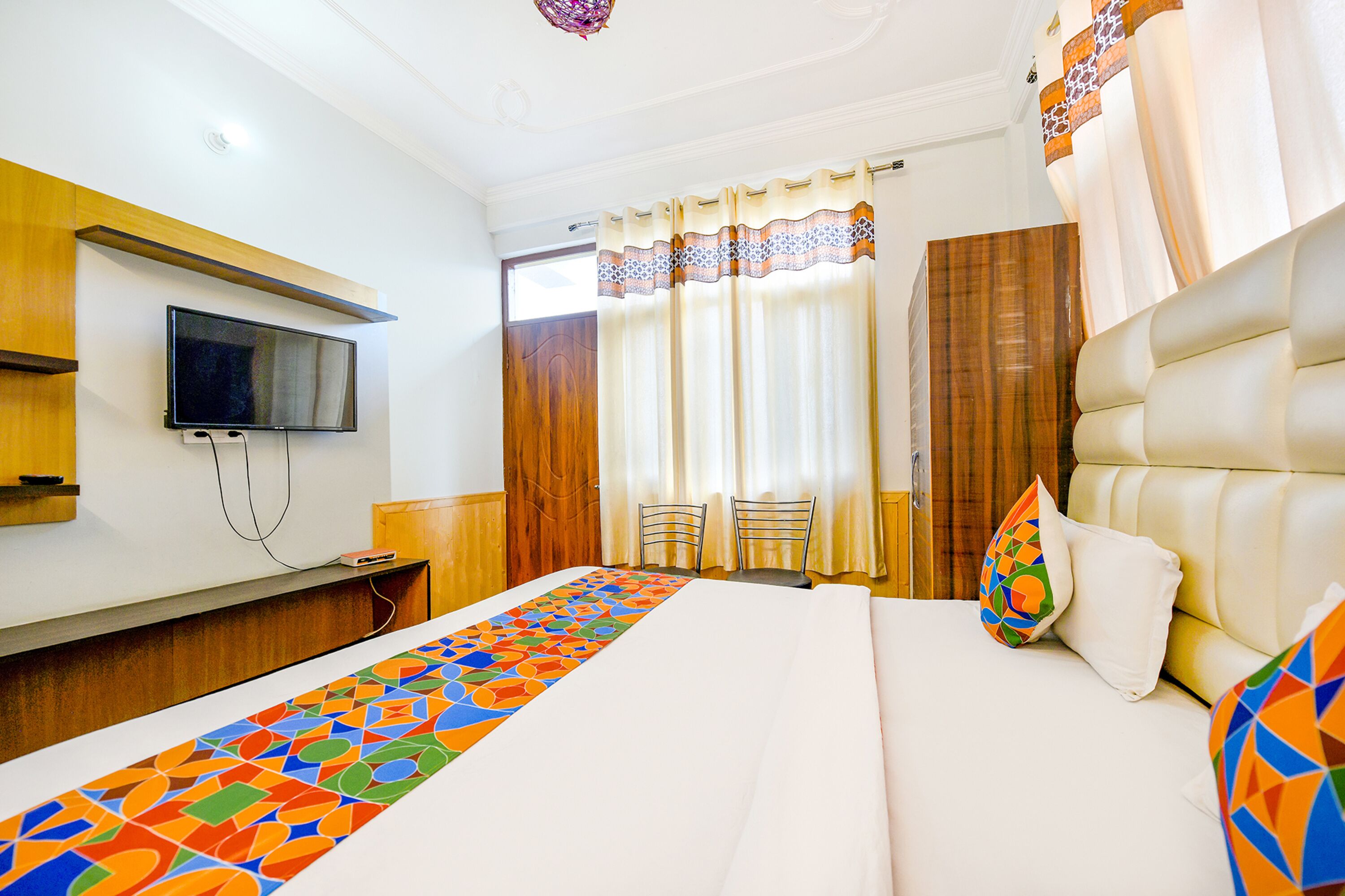 A hotel room with a bed, a TV mounted on the wall, a desk, and a window with curtains.