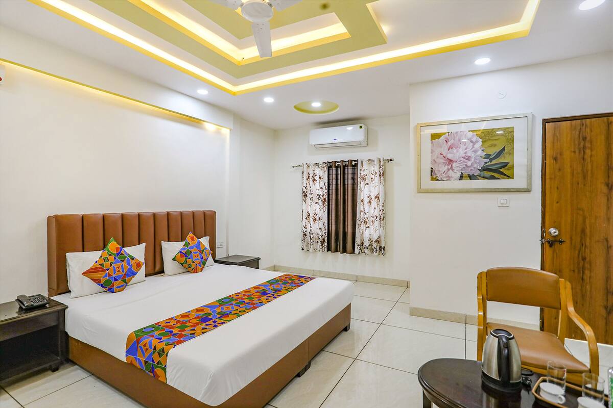 A hotel room with a large bed, a nightstand, a chair, a table with a kettle, and a painting on the wall.