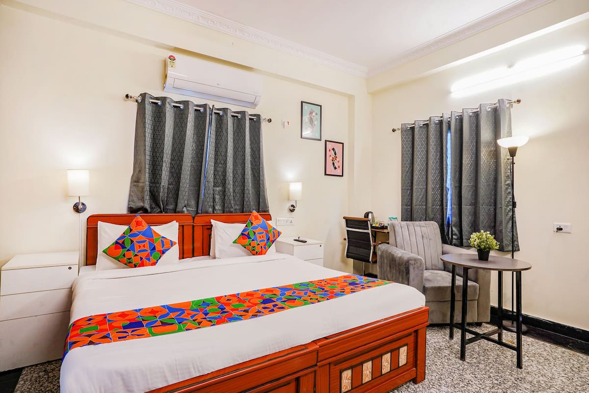Una habitación de hotel con una cama, una mesita pequeña, una silla, un equipo de aire acondicionado y una lámpara.