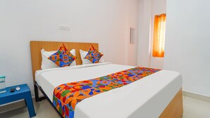 Una cama bien hecha con un edredón y almohadas de estampado geométrico colorido, una mesita de noche con un recipiente de vidrio y controles remotos, y una ventana con cortina naranja.