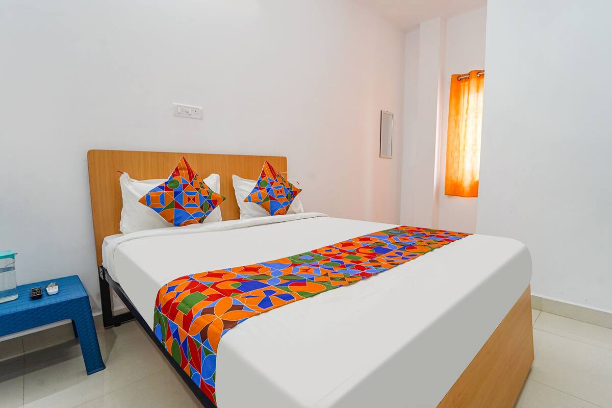 A neatly made bed with a colorful geometric-patterned bedspread and pillows, a small bedside table with a glass container and remote controls, and a window with an orange curtain.