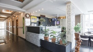 A hotel reception area with a modern design, including a reception desk, potted plants, and a wall-mounted clock. - Fabhotel Prime Suloka Towers (Kushalnagar)