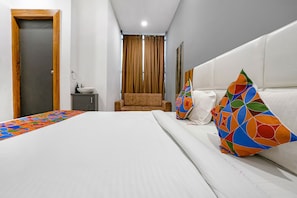A hotel room with a bed, a small bedside table, a chair, and a window with curtains. - Fabhotel Prime Dumas (Surat)