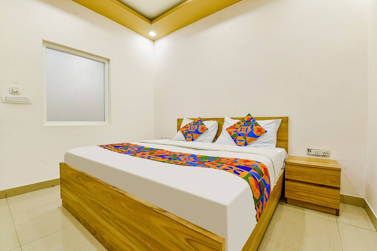 A bedroom with a wooden bed frame, a white mattress, and a colorful geometric-patterned bedspread. There is a wooden nightstand with a power outlet and a small drawer.