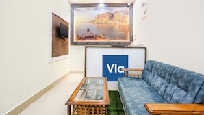 A living room with a sofa, a coffee table, a TV, and a wall-mounted painting. - Fabexpress Aditya Guest House (Varanasi)