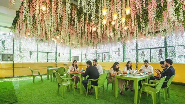 A group of people dining at a table with hanging floral decorations and a green artificial turf floor. - Fabexpress Aditya Guest House (Varanasi)