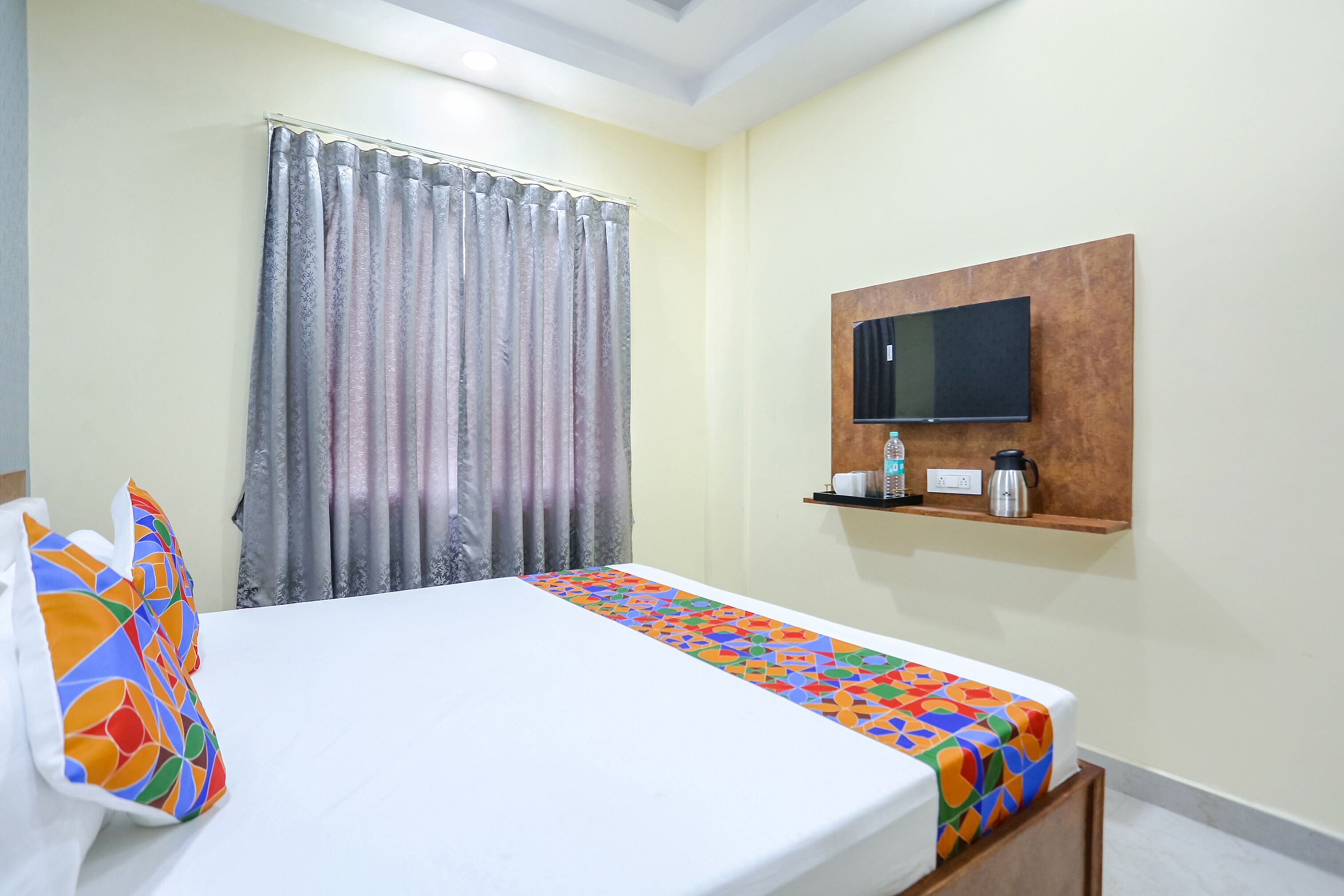 A hotel room with a bed, a TV mounted on the wall, a shelf with a kettle and other items, and a window with curtains.