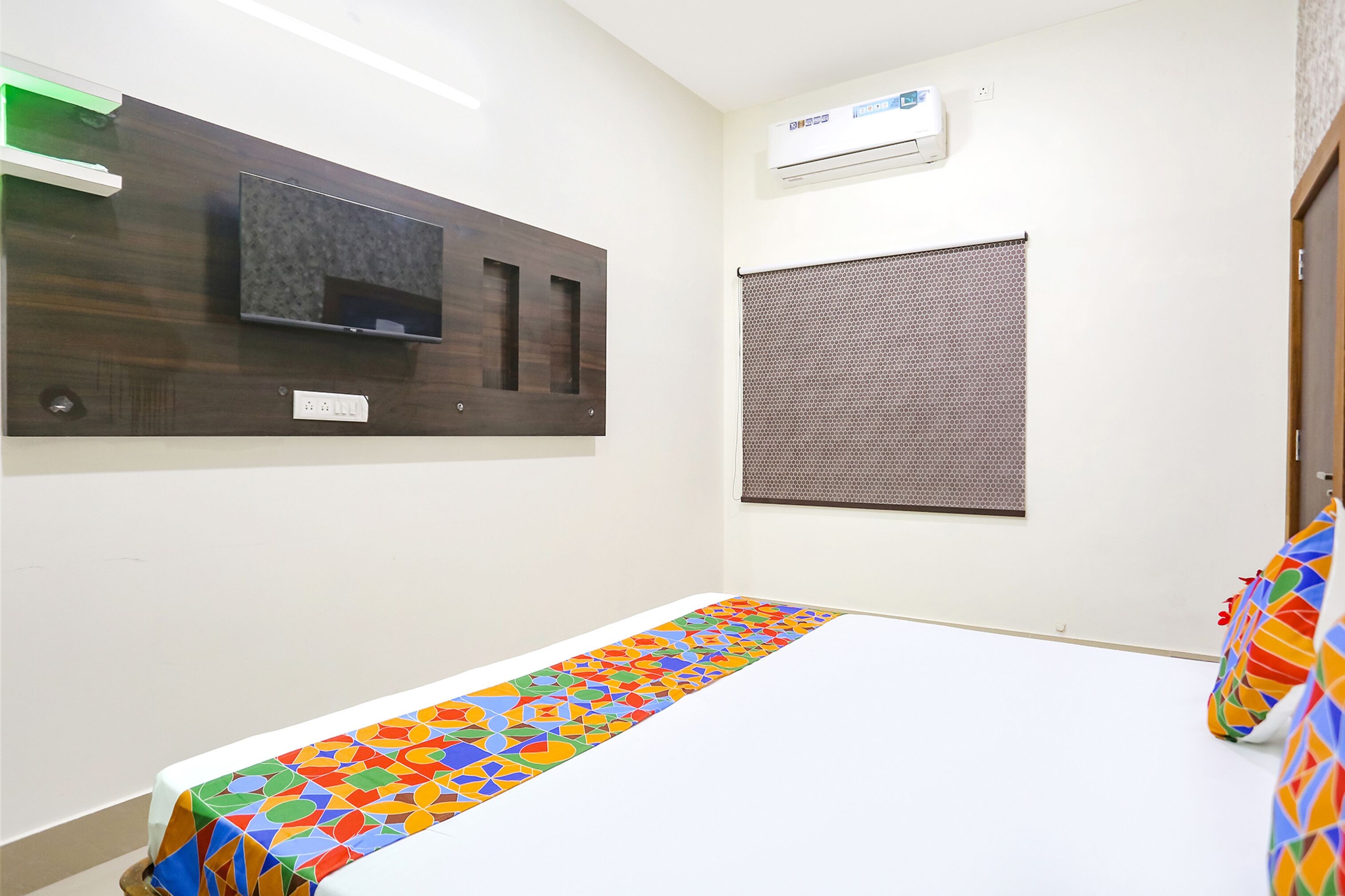 Una habitación pequeña y limpia con una cama, televisión montada en la pared, aire acondicionado y ventana con persianas.