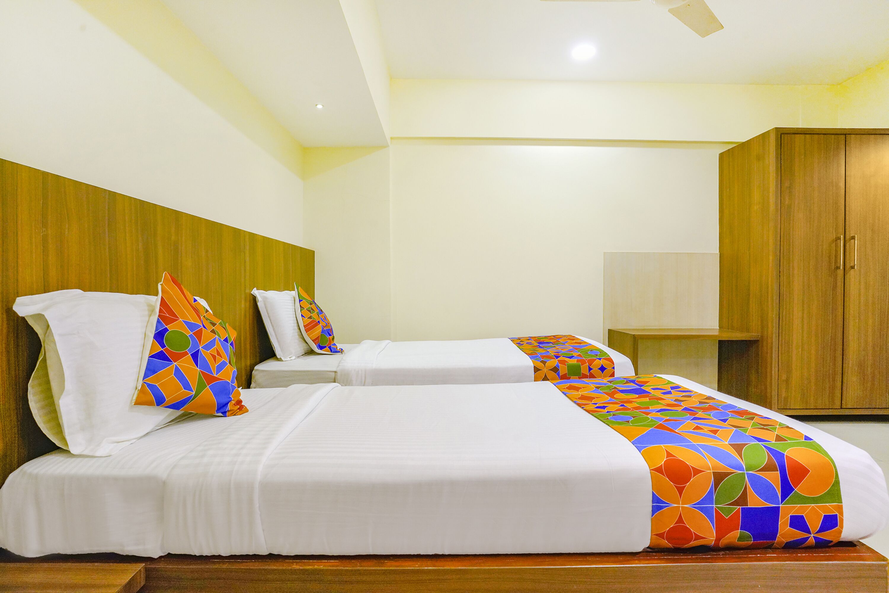 Une chambre d’hôtel avec deux lits, une tête de lit en bois, du linge de lit blanc et une couvre-lit aux motifs géométriques et aux couleurs vives.