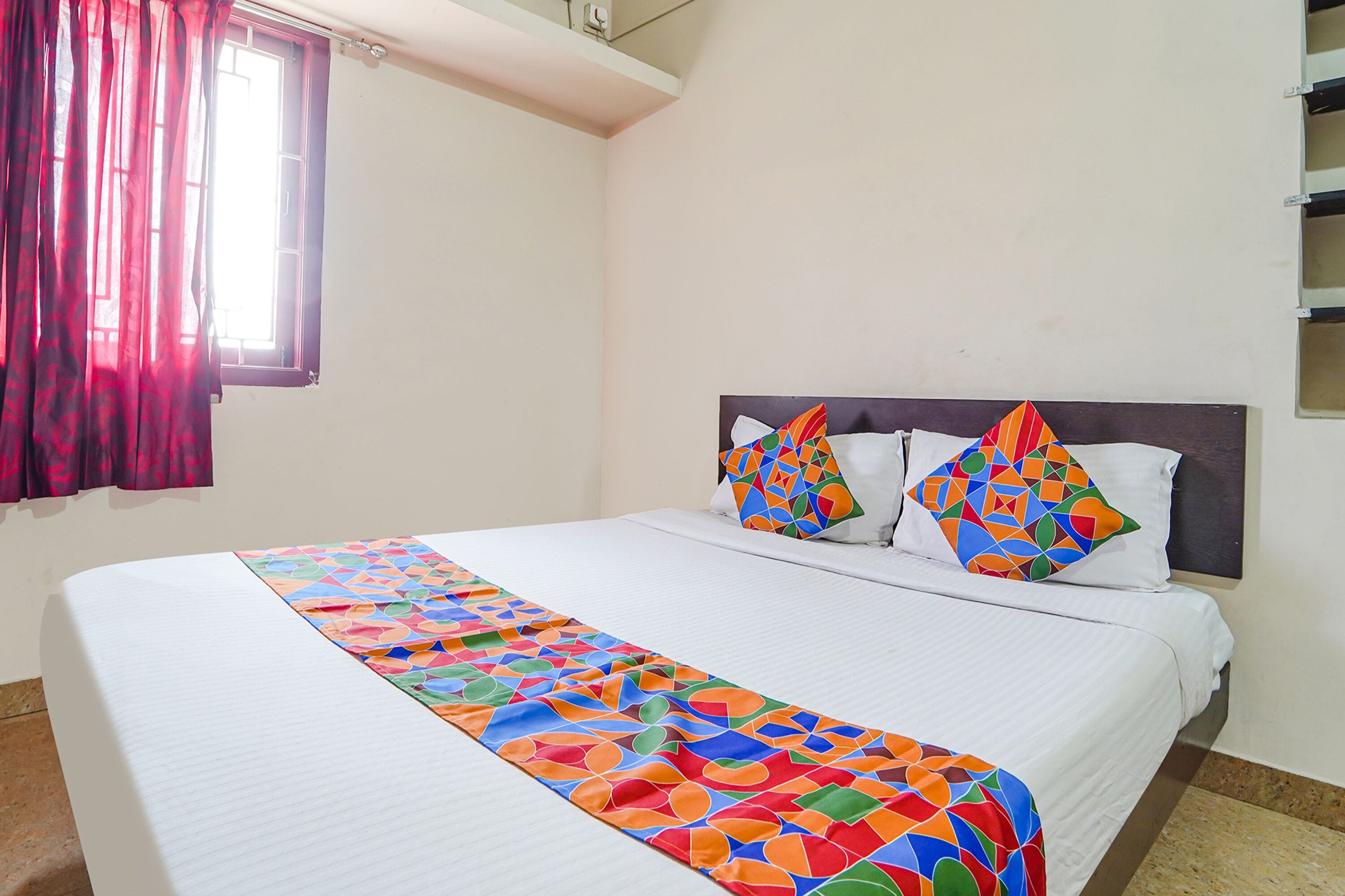 A neatly made bed with a colorful geometric-patterned bedspread, two decorative pillows, and a window with red curtains.
