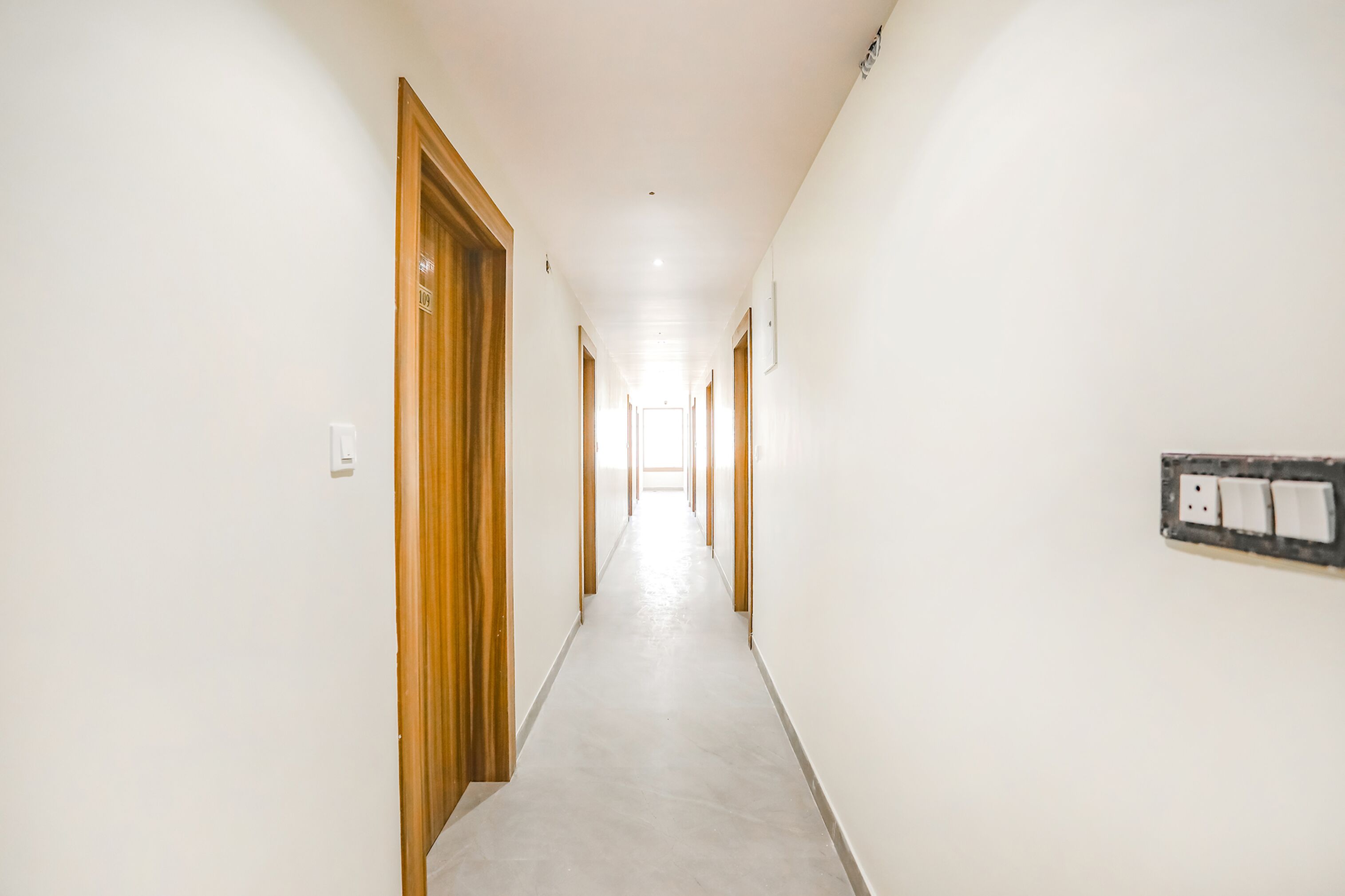 A long, narrow hallway with wooden doors on one side and a light switch on the other.