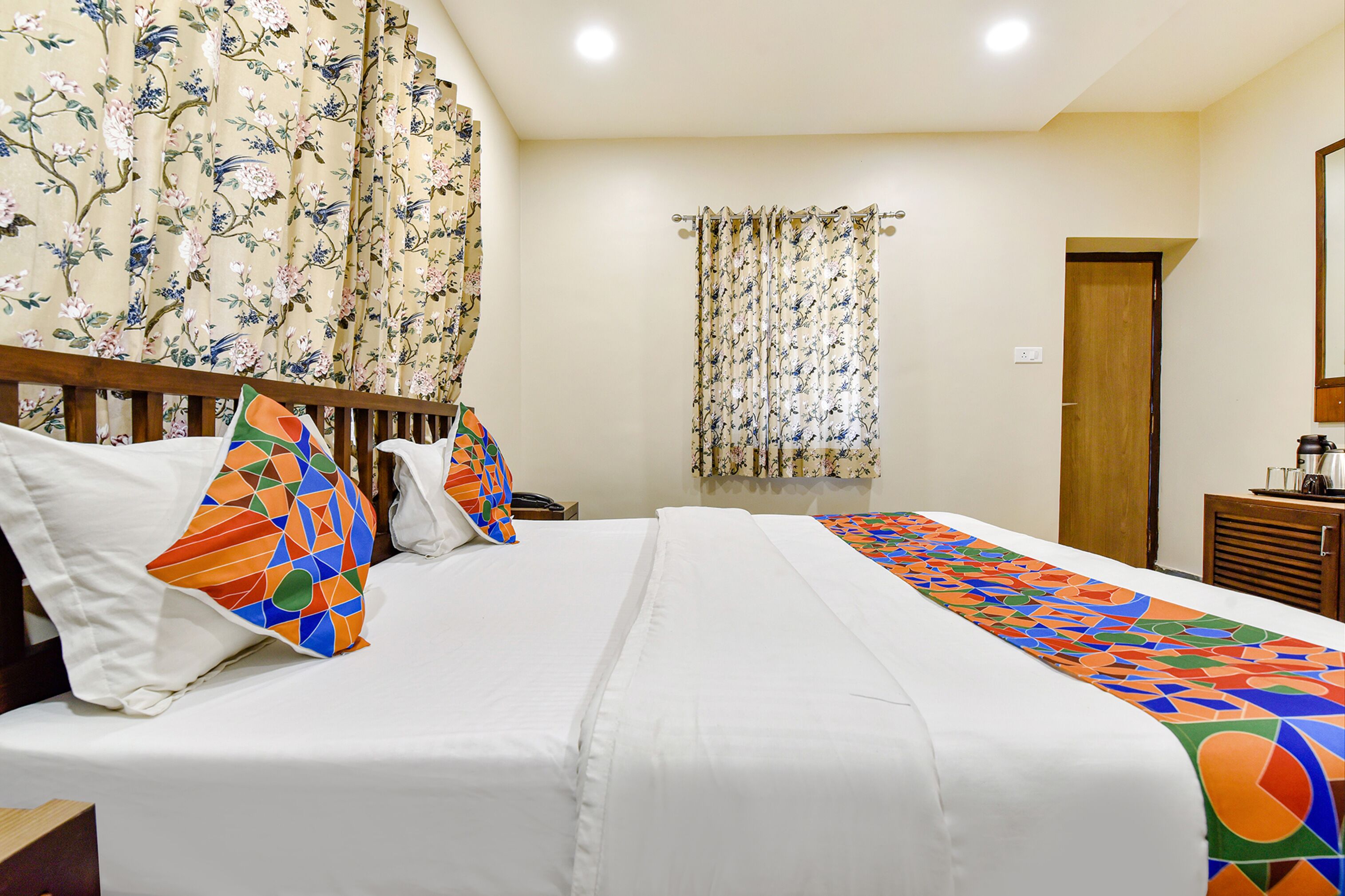 A hotel room with a large bed, floral curtains, a wooden headboard, and a small wooden side table with a kettle.