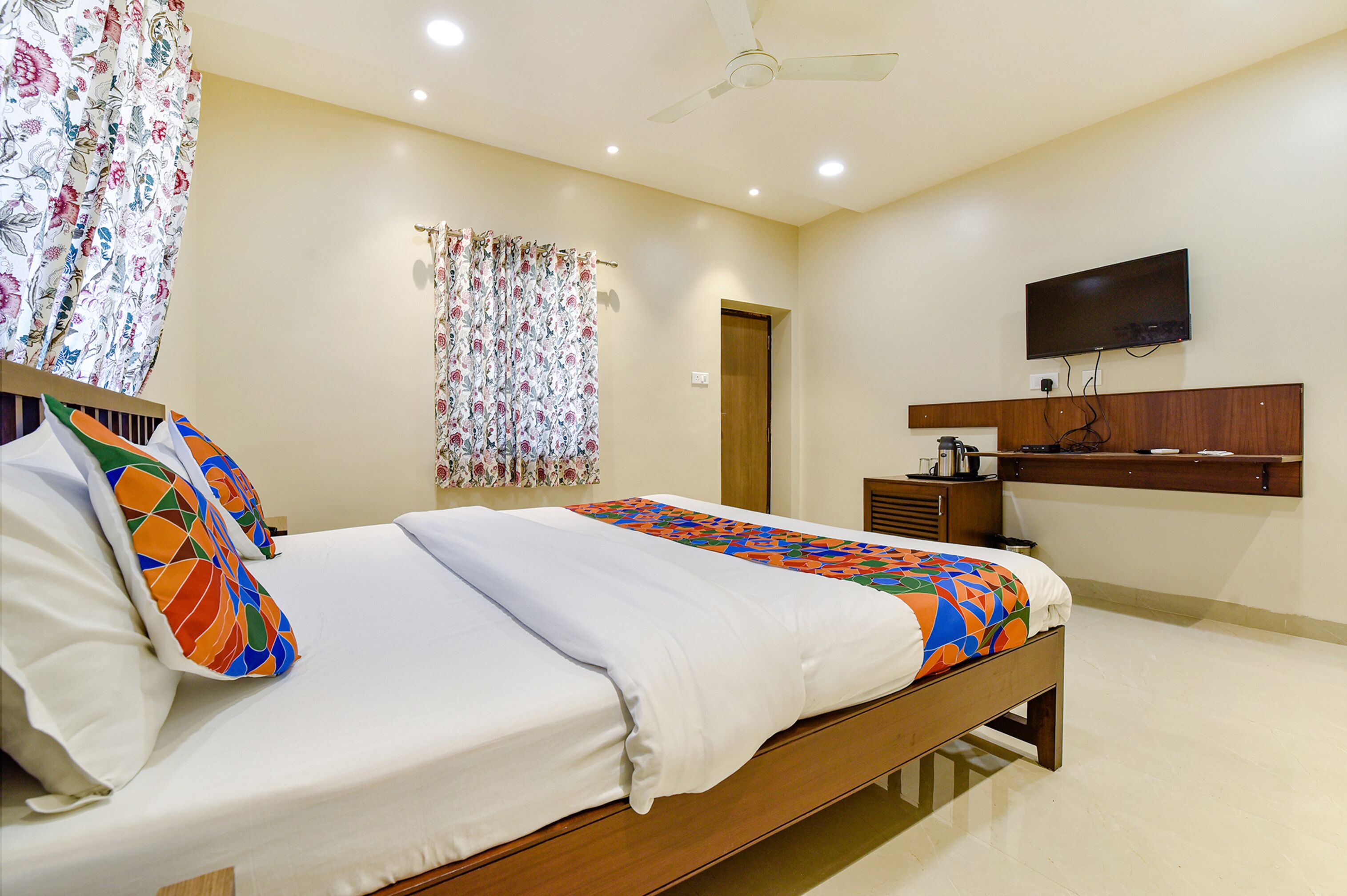 A neatly made bed with colorful pillows, a television mounted on the wall, and a ceiling fan.