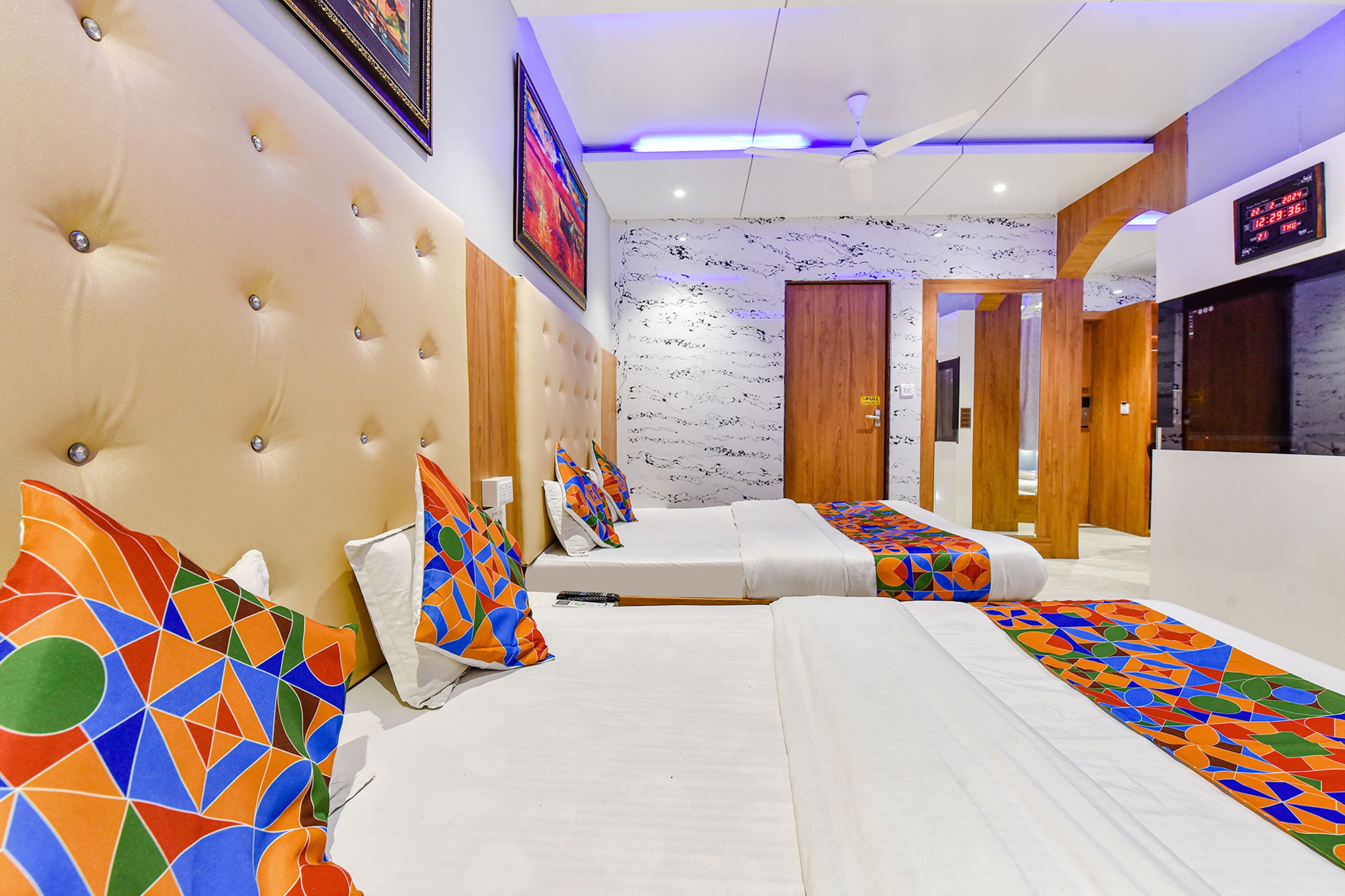 A hotel room with a tufted headboard, a bed with colorful geometric-patterned bedding, a white wall with a black and white abstract design, and a digital clock displaying the time.
